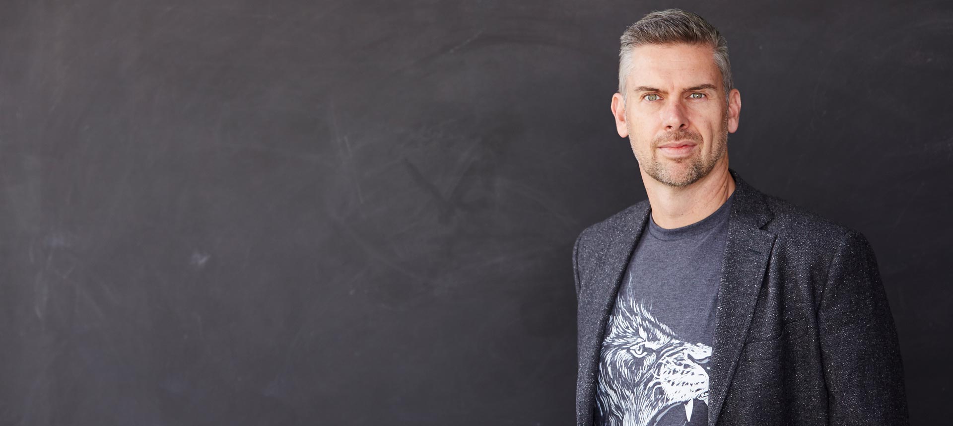 Mark Dobson standing against a blackboard wall, wearing a suit jacket and a lion print t-shirt